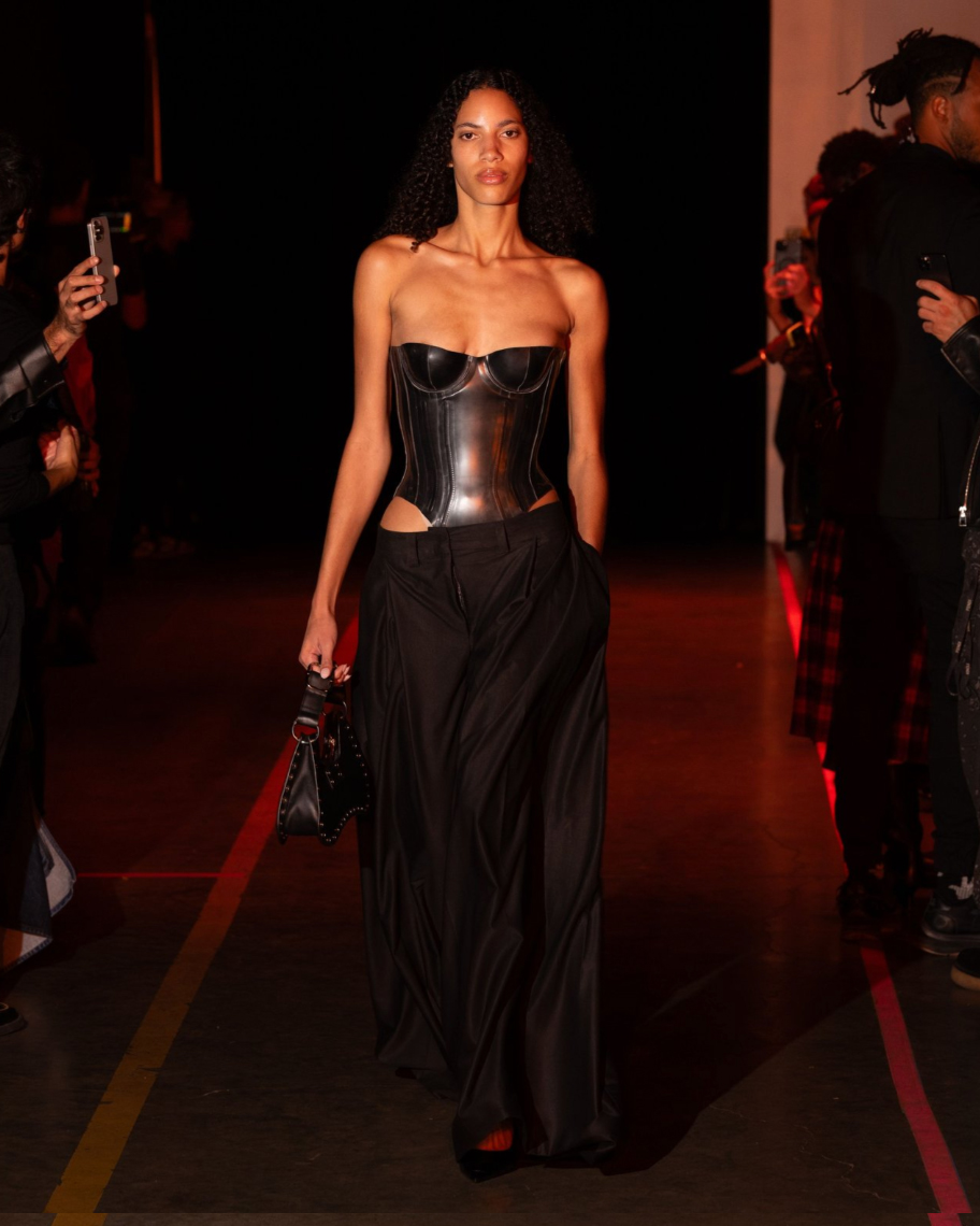 Slender female model with tan skin wearing a latex corset and black pants walking on a runway against a dark backdrop